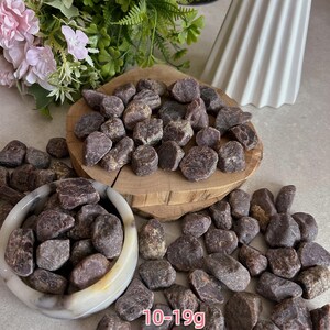 Peut inclure: Une collection de pierres de grenat brutes, brun rouge&acirc;tre fonc&eacute;, avec des pi&egrave;ces pesant entre 10 et 19g. Les pierres sont pr&eacute;sent&eacute;es dans un petit bol en pierre de couleur claire et empil&eacute;es sur une tranche de bois circulaire &agrave; bord naturel.