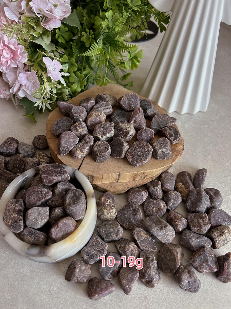 Peut inclure: Une collection de pierres de grenat brutes, brun rouge&acirc;tre fonc&eacute;, avec des pi&egrave;ces pesant entre 10 et 19g. Les pierres sont pr&eacute;sent&eacute;es dans un petit bol en pierre de couleur claire et empil&eacute;es sur une tranche de bois circulaire &agrave; bord naturel.