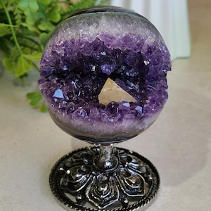 Puede incluir: Una geoda de amatista esférica, que muestra un vibrante interior de cristal púrpura y una formación de cristal de color claro. La geoda está montada sobre un soporte decorativo de metal plateado con una base ornamentada, creando una pieza de exhibición llamativa.