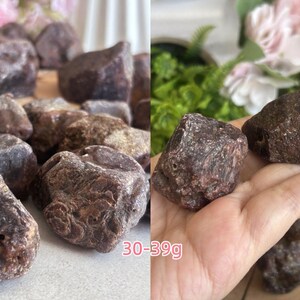Peut inclure: Une collection de pierres de grenat brutes, brun rouge&acirc;tre fonc&eacute;, aux surfaces irr&eacute;guli&egrave;res et facett&eacute;es avec un aspect textur&eacute;. Les pierres varient en forme et en taille, certaines pi&egrave;ces &eacute;tant clairement &eacute;tiquet&eacute;es comme pesant entre 30-39g.