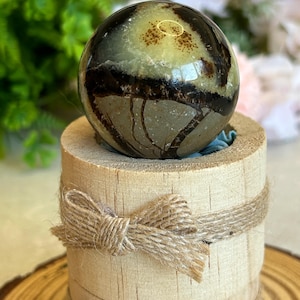 Puede incluir: Una piedra esférica pulida con una mezcla de patrones en verde pálido y marrón oscuro, que descansa sobre un soporte cilíndrico de madera clara. El soporte está decorado con dos vueltas horizontales de cuerda de yute natural.