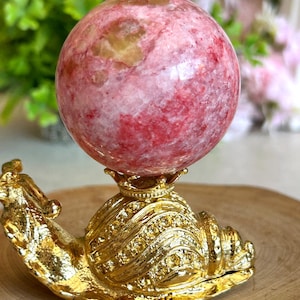 May include: A polished pink and red mottled stone sphere resting on a gold-colored metal snail-shaped stand. The snail stand features a textured, shell-like back with a small crown-shaped holder at the top to support the spherical stone.
