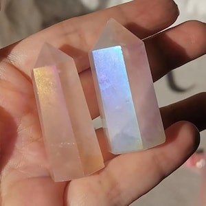 Puede incluir: Dos puntas de cristal de cuarzo iridiscente. Los cristales tienen forma rectangular con una parte superior puntiaguda. Uno tiene un tono dorado, el otro un brillo azul y morado. Los cristales se sostienen en una mano.