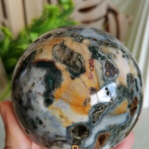 Puede incluir: Una esfera de piedra preciosa pulida con un patrón moteado de tonos grises, marrones, verdes y naranjas. El orbe se sostiene en una mano, mostrando su forma lisa y redondeada. La superficie de la esfera tiene una apariencia natural y terrosa.
