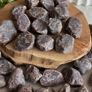 Puede incluir: Una colección de piedras de granate en bruto, sin pulir, de color granate oscuro y rojo parduzco, con formas irregulares y dentadas y superficies texturizadas. Estos especímenes minerales naturales están esparcidos sobre una base de madera.