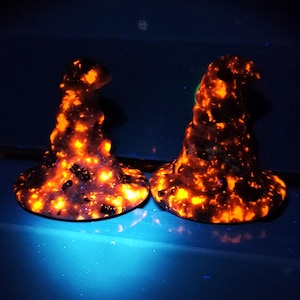 Può includere: Due sculture luminose, arancioni e nere che assomigliano a cappelli da strega. I cappelli hanno una superficie strutturata con macchie scure e un bagliore brillante e infuocato. I cappelli sono esposti su una superficie blu.