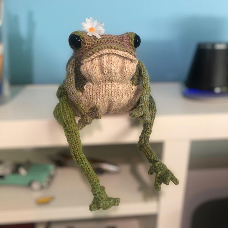 May include: A handmade knitted frog doll with large black eyes and a small white daisy on its head. The frog is primarily green and tan, with long, slender legs and webbed feet. The frog is sitting on a white surface.