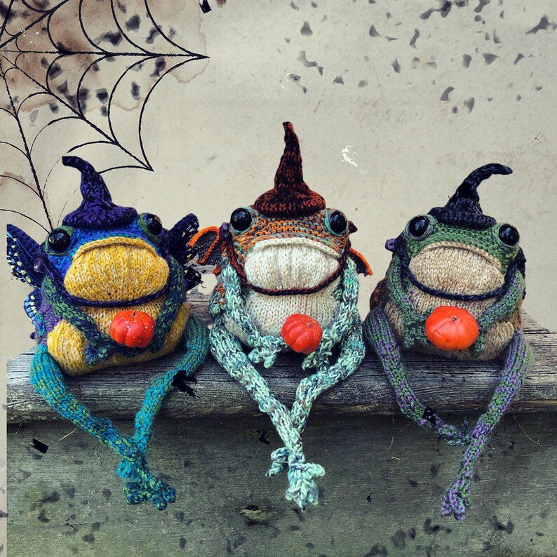 May include: Three whimsical, knitted frog dolls with witch hats and small orange pumpkins. The frogs are in shades of blue, green, and orange, with detailed features and long, dangling legs. A spider web design is in the background.