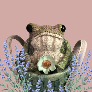May include: A knitted frog doll with large black eyes sits in a green woven basket, holding a white and orange flower. Lavender sprigs frame the basket against a pink background. The frog is primarily beige and green.
