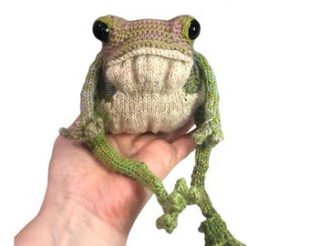 May include: A knitted frog toy with large black eyes, in shades of green, tan, and pink. The frog has long, detailed legs and the text "KNITTING PATTERN" is visible at the bottom. The MageWood Whimsies logo is in the top right corner.