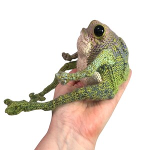 May include: A handmade, knitted frog figurine with large black eyes. The frog is primarily green with accents of purple and tan. The frog is sitting with its legs extended. The frog is a unique, whimsical piece.