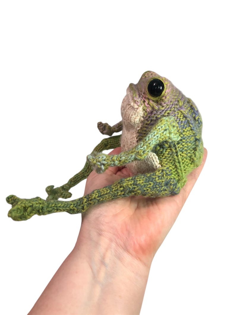 May include: A handmade, knitted frog figurine with large black eyes. The frog is primarily green with accents of purple and tan. The frog is sitting with its legs extended. The frog is a unique, whimsical piece.