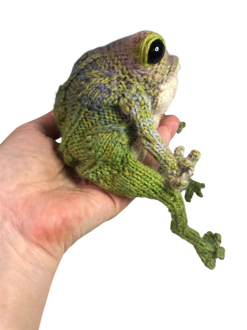 May include: A knitted frog figurine in shades of green, purple, and cream. The frog has large black eyes and is posed with its legs extended. The handmade amphibian is held in a hand against a white background.