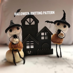 May include: A Halloween knitting pattern featuring two whimsical witch dolls with white hair, black hats, and pumpkin baskets. They stand near a black wooden haunted house. The dolls are perched on a white pumpkin.