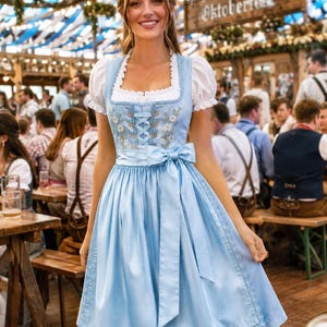 May include: A light blue dirndl dress featuring a white puff-sleeve blouse, a square neckline, and a bodice with floral embroidery and lace-up detailing. The skirt includes a matching satin ribbon waist tie and a delicate lace trim at the hem.