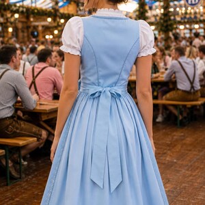 May include: A light blue dirndl dress featuring a white blouse with short puffed sleeves, a high neckline, and a fitted bodice. The skirt is knee-length with a decorative lace trim along the hem and a matching blue fabric tie at the waist.