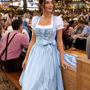 May include: A light blue dirndl dress featuring a white blouse with puffed sleeves, a square neckline, and a bodice with lace-up detailing. The dress includes a matching apron with a tied bow at the waist and delicate floral embroidery on the front.