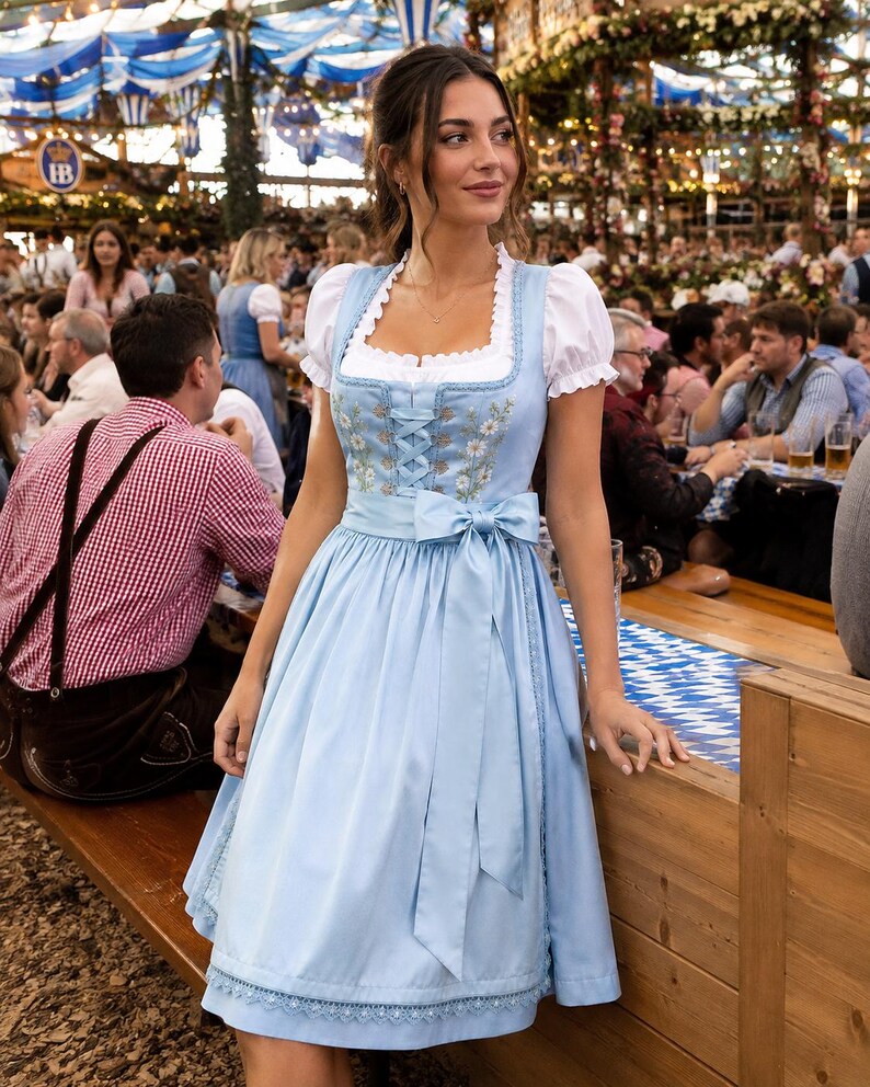 May include: A light blue dirndl dress featuring a white blouse with puffed sleeves, a square neckline, and a bodice with lace-up detailing. The dress includes a matching apron with a tied bow at the waist and delicate floral embroidery on the front.
