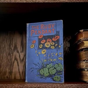 May include: An antique book, "The Ruby Pendant", with a blue cover and floral artwork in orange, yellow, and green. It sits on a wooden shelf beside a stack of old books, suggesting a literary theme.