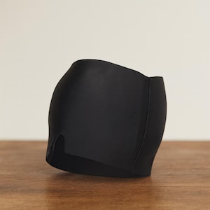 Op de afbeelding: Een zwart, nauwsluitend kledingstuk, mogelijk badkleding of lingerie, met een gladde textuur. Het item heeft een gebogen vorm en een klein spleetdetail aan de voorkant. Het ligt op een houten oppervlak.