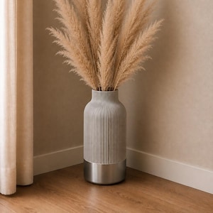 Könnte beinhalten: Eine hohe, zylindrische Vase mit einem gerippten, hellgrauen Oberteil und einem glatten, metallisch silbernen Boden. Die Vase ist mit einem großen Arrangement aus hohen, federartigen, beigen Pampasgraswedeln gefüllt, die aufrecht stehen.