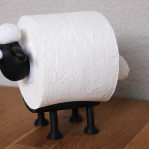 May include: A black sheep-shaped toilet paper holder featuring a black head with white eyes and a white tuft of wool on top. The body is formed by a roll of white toilet paper with a floral embossed pattern, supported by four black legs.