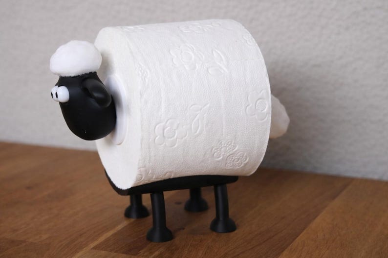 May include: A black sheep-shaped toilet paper holder featuring a black head with white eyes and a white tuft of wool on top. The body is formed by a roll of white toilet paper with a floral embossed pattern, supported by four black legs.