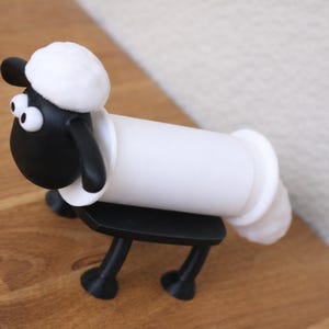 May include: A black and white sheep-shaped toilet paper roll holder featuring a cartoon-style head with large white eyes, a fluffy white top, and black ears. The body consists of a horizontal white cylinder supported by four black legs and a base.
