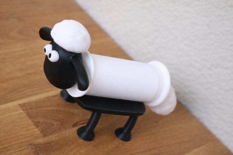 May include: A black and white sheep-shaped toilet paper roll holder featuring a cartoon-style head with large white eyes, a fluffy white top, and black ears. The body consists of a horizontal white cylinder supported by four black legs and a base.