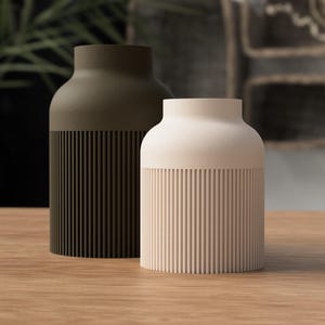 Könnte beinhalten: Zwei zylindrische Vasen mit einem glatten oberen Teil und einem vertikal gerippten unteren Körper. Eine Vase ist dunkel olivgrün, die andere ist cremefarben. Beide verfügen über eine weite Öffnung an der Oberseite und einen flachen Boden.