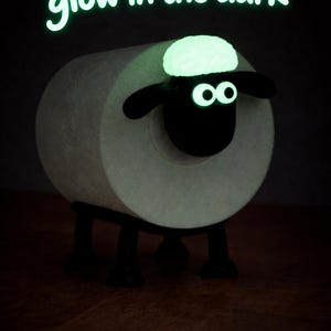 May include: A toilet paper roll holder shaped like a sheep with a black head and legs, featuring glow-in-the-dark eyes and a tuft of wool on its head. The text "Variante glow in the dark" is displayed in glowing green letters above the sheep figure.