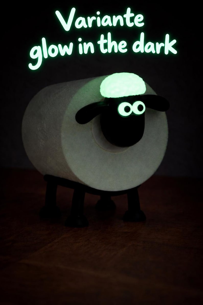 May include: A toilet paper roll holder shaped like a sheep with a black head and legs, featuring glow-in-the-dark eyes and a tuft of wool on its head. The text "Variante glow in the dark" is displayed in glowing green letters above the sheep figure.