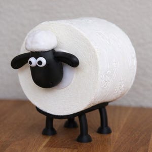 May include: A white toilet paper roll holder shaped like a sheep. The holder features a black sheep head with white googly eyes and a white tuft of wool on top, attached to the side of the roll. It has four black legs supporting the roll.