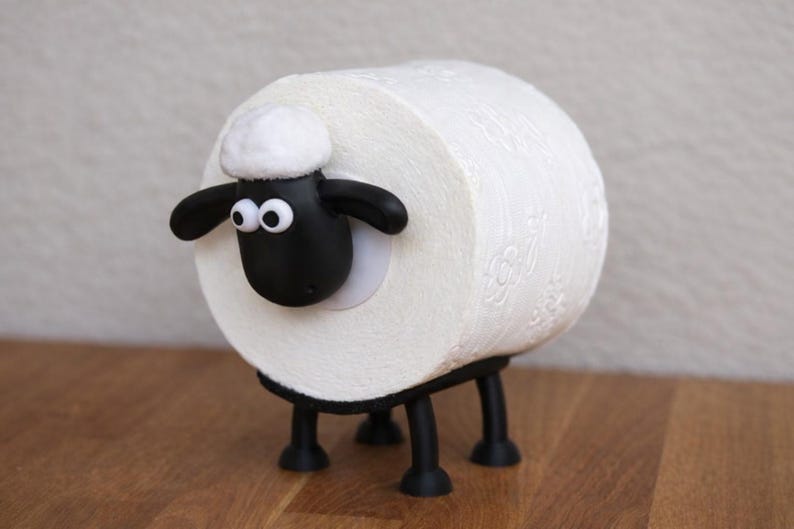 May include: A white toilet paper roll holder shaped like a sheep. The holder features a black sheep head with white googly eyes and a white tuft of wool on top, attached to the side of the roll. It has four black legs supporting the roll.