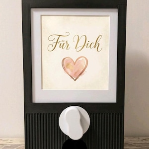May include: A black wall-mounted dispenser featuring a vertical ribbed texture on the lower section and a white dispensing knob. The upper portion displays a framed print with the text "Für Dich" in gold script above a watercolor heart.