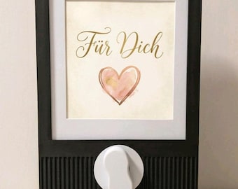 May include: A black wall-mounted dispenser featuring a vertical ribbed texture on the lower section and a white dispensing knob. The upper portion displays a framed print with the text "Für Dich" in gold script above a watercolor heart.