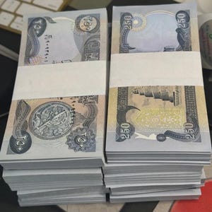 May include: Two stacks of banknotes, each secured with a white paper band. The light blue currency features detailed patterns and Arabic script. The value of 250 is printed on each note. The stacks are on a desk, with a keyboard visible in the background.