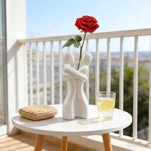 May include: A white ceramic vase in the shape of two embracing figures holds a single red rose. The vase sits on a white table with a woven coaster and a glass of water with lemon slices. The background shows a balcony railing and a sunny sky.