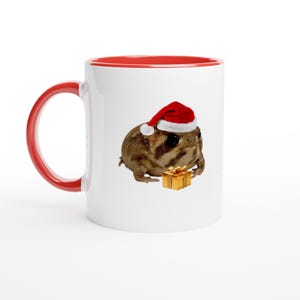 May include: White ceramic mug with a red interior and handle. The mug features a brown frog wearing a Santa hat and holding a small gold gift box. A festive holiday-themed mug.