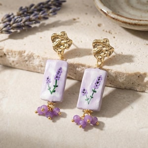 Può includere: Orecchini dorati con un pendente rettangolare color lavanda con un rametto di lavanda dipinto a mano. Gli orecchini hanno una parte superiore dorata strutturata e un gruppo di perline viola nella parte inferiore. È presente anche un rametto di lavanda.