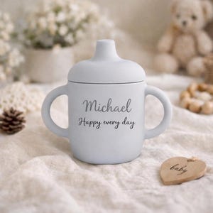 May include: A light blue baby sippy cup with two handles and a lid. The cup has the name "Michael" and the phrase "Happy every day" printed on it. A wooden heart-shaped tag that says "baby" is next to the cup.
