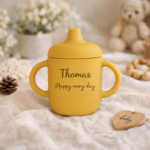 May include: A yellow sippy cup with two handles and a lid. The cup has the name "Thomas" and the phrase "Happy every day" printed on the front. A wooden heart-shaped tag with the word "baby" is next to the cup.