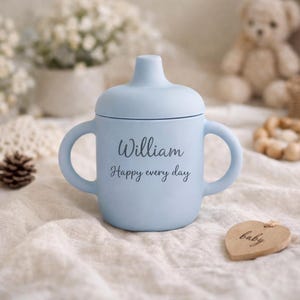 May include: A light blue baby sippy cup with two handles and a lid. The cup has the name "William" and the phrase "Happy every day" printed on the front. A wooden heart-shaped tag with the word "baby" is also visible.