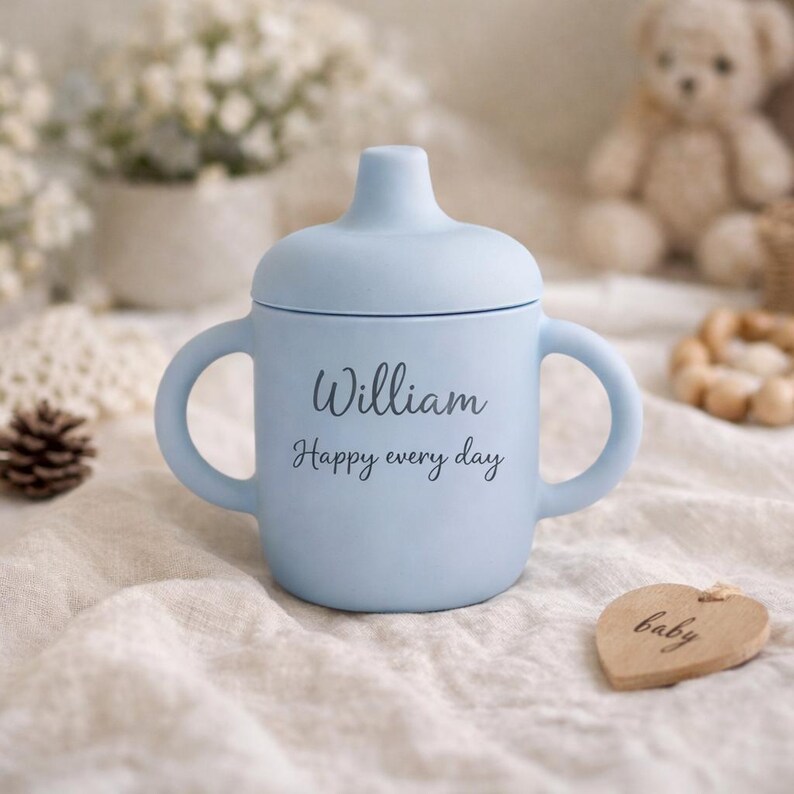 May include: A light blue baby sippy cup with two handles and a lid. The cup has the name "William" and the phrase "Happy every day" printed on the front. A wooden heart-shaped tag with the word "baby" is also visible.