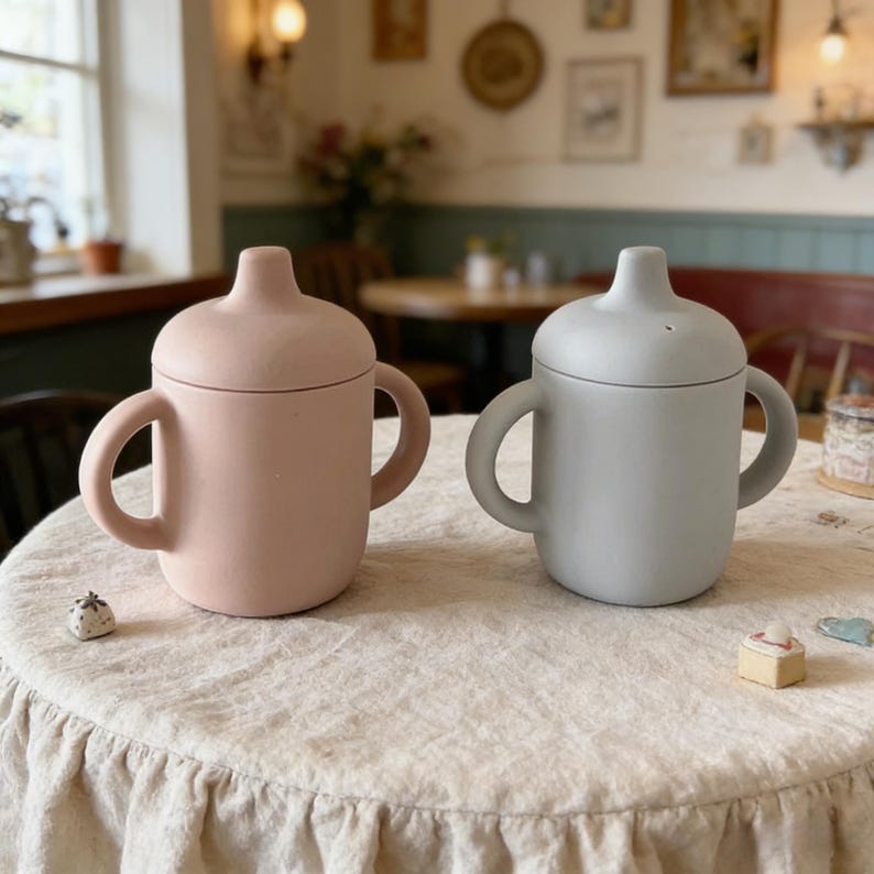 May include: Two silicone baby cups with handles and lids. One cup is a soft pink, the other is gray. Both cups are on a cream-colored tablecloth. The cups have a rounded shape and a small spout on the lid.