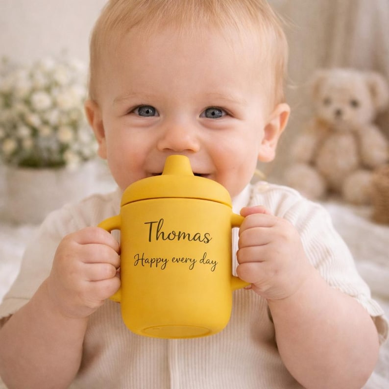 May include: A smiling baby holds a yellow sippy cup with handles. The cup has the name "Thomas" and the phrase "Happy every day" printed on it in black. A teddy bear is in the background.