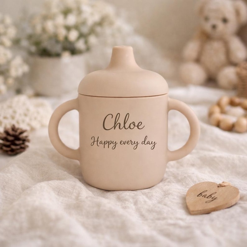 May include: A beige sippy cup with a lid and two handles. The cup has the name "Chloe" and the phrase "Happy every day" printed on it. A wooden heart-shaped tag with the word "baby" is also visible.