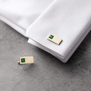 May include: A pair of rectangular gold-tone cufflinks, each featuring a single square-cut emerald green stone set on one side. One cufflink is attached to the corner of a folded white textured fabric cuff, while the other rests nearby.