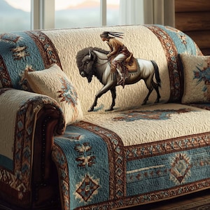 May include: A sofa with a cream-colored quilted cover featuring a Native American riding a bison. The cover has blue and brown geometric patterns along the edges. Matching throw pillows with similar designs complete the look.