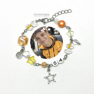 Könnte beinhalten: Ein silberfarbenes Kettenarmband mit einem runden Fotoanhänger einer Person in einem gelb-schwarzen Kapuzenpullover, verziert mit orangefarbenen, gelben und klaren Perlen, sternförmigen Anhängern, einer Spirale und weißen Perlen mit der Aufschrift 04.